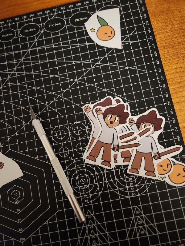 A picture of a some cut stickers depicting my original character Mac (a small child with a wooden sword in a victory pose). Other cut sickers depict an orange winking. Near the stickers there's a precision knife and a cutting surface.